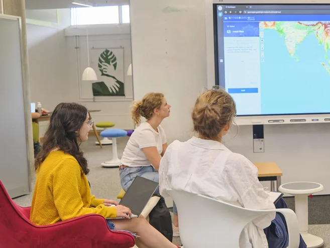 Frau präsentiert Weltkarte auf großem Bildschirm im Digital Creative Space, vier Personen sitzen und hören zu