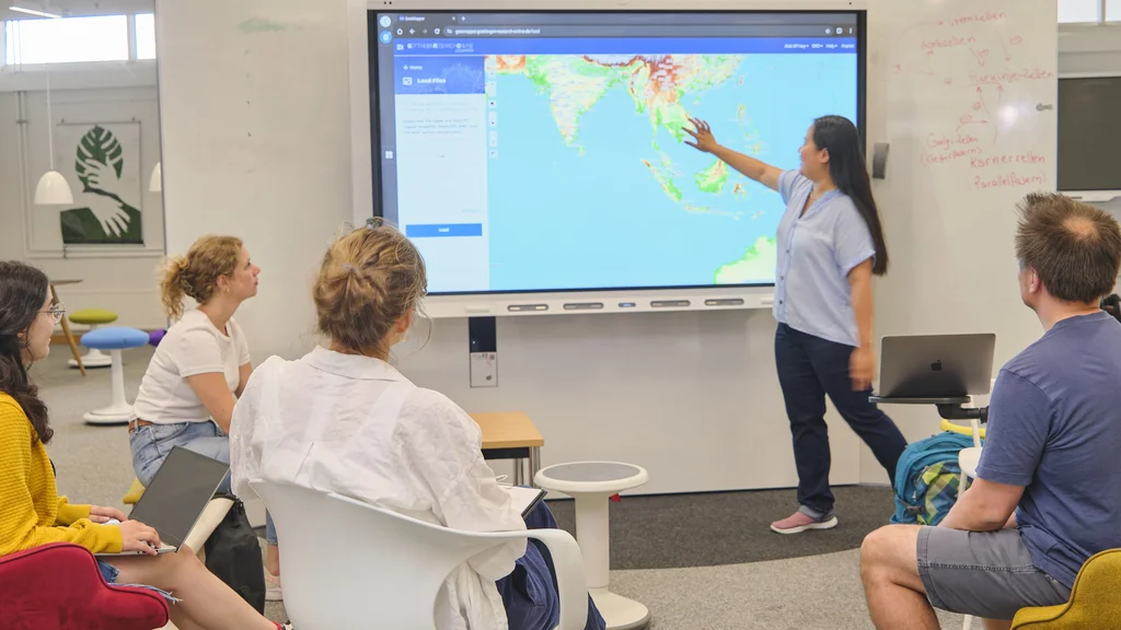 Frau präsentiert Weltkarte auf großem Bildschirm im Digital Creative Space, vier Personen sitzen und hören zu