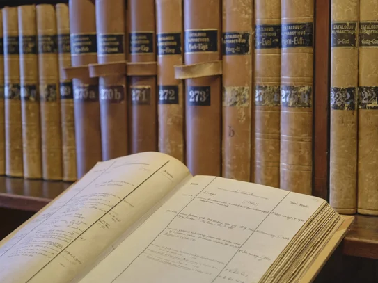 Im Hintergrund Bibliothekskataloge auf einem Regal, im linken vorderen Bereich ein aufgeschlagener Bibliothekskatalog