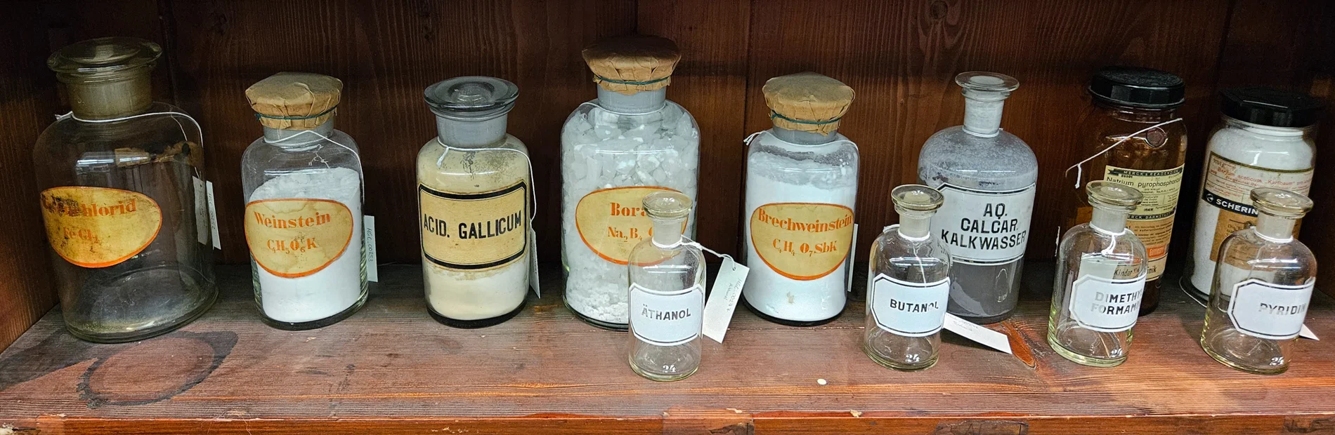 Holzregal mit zehn alten Apothekergläsern verschiedener Größen, gefüllt mit weißen Pulvern, beschriftet mit chemischen Substanzen