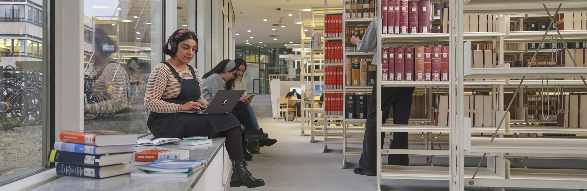Zwei Frauen sitzen in der Bibliothek auf einer Fensterbank, eine arbeitet an einem Laptop, im Hintergrund Bücherregale und weitere Personen