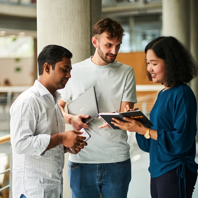 Drei Personen in der Zentralbibliothek. Eine Person hält ein Tablet, eine andere ein Laptop. Die dritte Person hat ein Smartphone in der Hand.