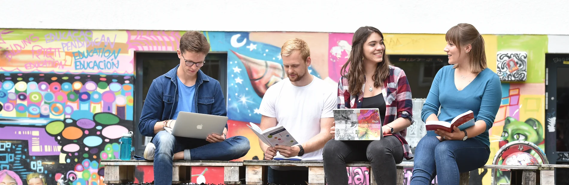 Junge Menschen sitzen auf einer Bank vor einem Gebaeude mit bunten Graffiti