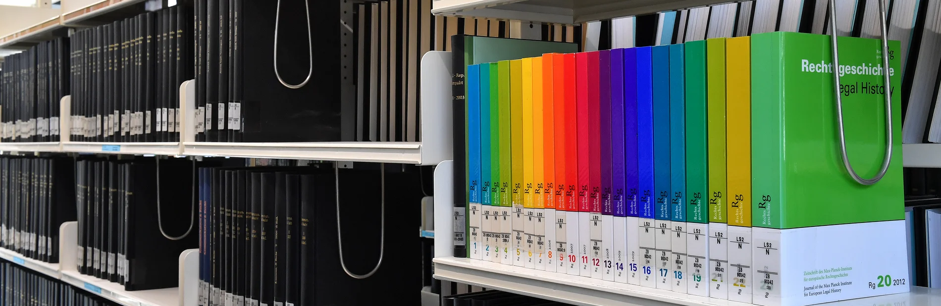 In einem Regal stehen bunte Buchrücken in den Regenbogenfarben umgeben von dunklen Büchern