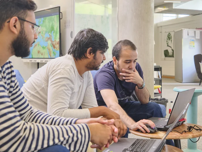 Drei Personen sitzen im Digital Creative Space an einem Tisch, zwei nutzen Laptops, eine Person zeigt auf den Bildschirm