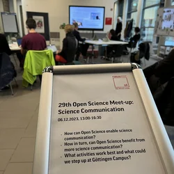 Hinweisschild mit Beschreibung zum Open Science Meetup mit Meetingraum und Bildschirm im Hintergrund
