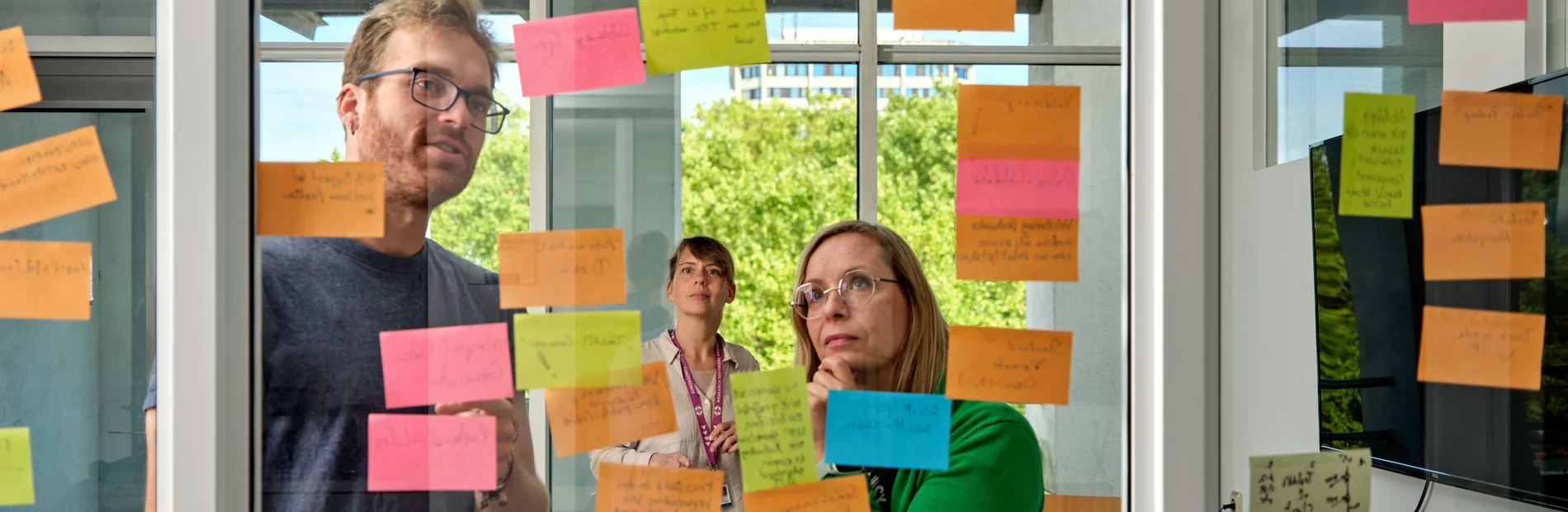 Personen stehen vor einer Glaswand mit beschriebenen Notizzetteln und beraten sich.