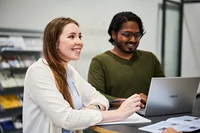 Zwei Personen stehen an einem Tisch, darauf steht ein Laptop. Eine Person hört aufmerksam zu
